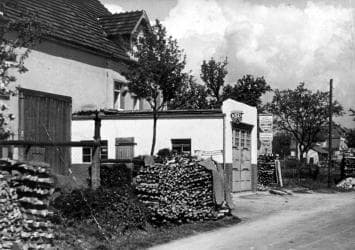 Historisches Schwarz-Weiß-Foto: das erste Werkstattgebäude von Rüdinger in Aglasterhausen mit Holzstapeln davor